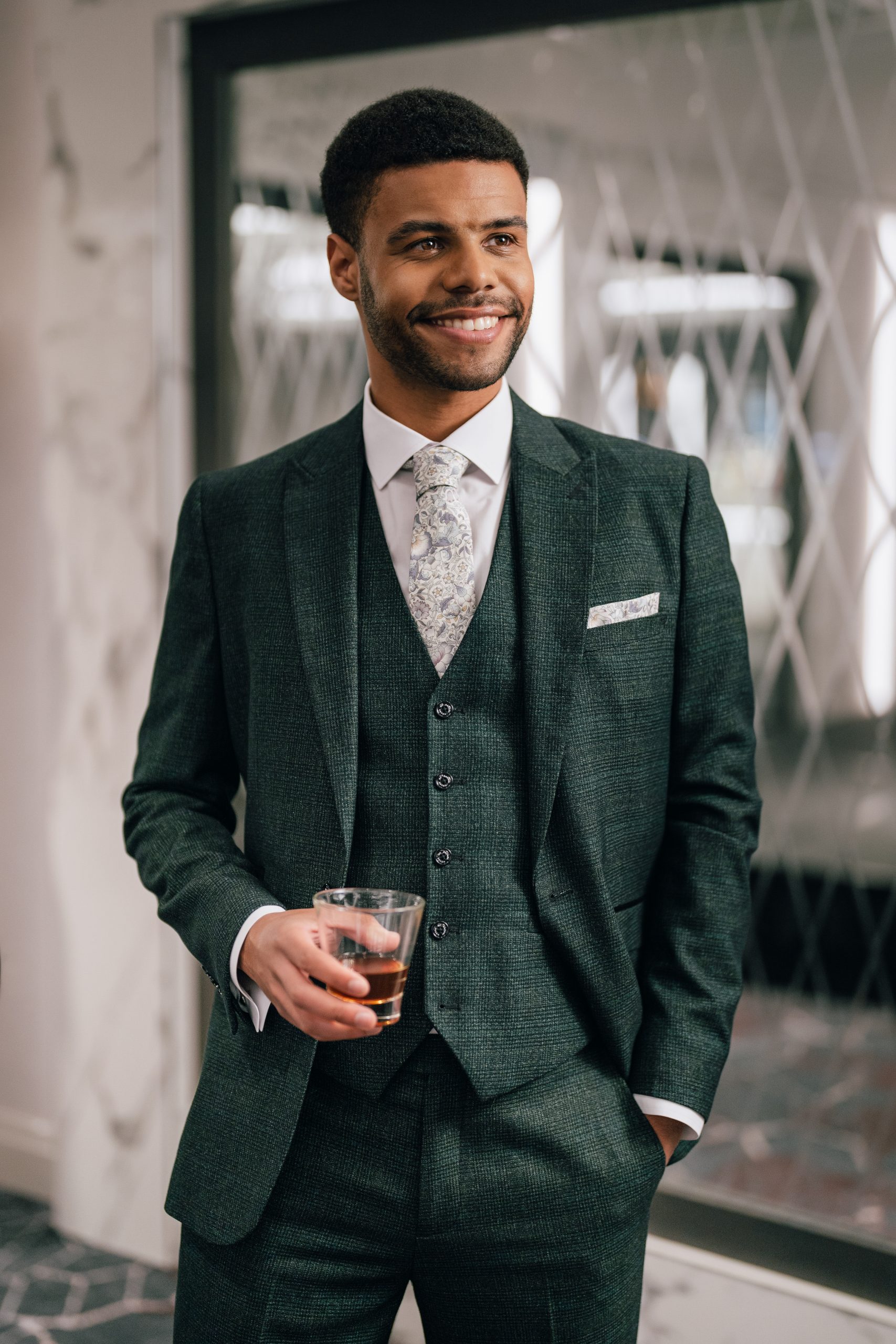 Model stands with whiskey glass wearing drak green cavani suit from ACS rental against mirrored glass background